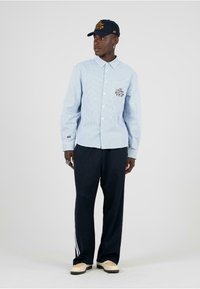 Himmelblau gestreifte Baumwollhemd mit einer Tasche; kombiniert mit dunklen Hosen mit weißen seitlichen Streifen. Getragen mit einer dunklen Mütze und hellen Schuhen.