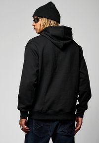 Hoodie noir en coton, doté d'une poche kangourou à l'avant et d'une capuche avec cordon de serrage, porté avec un jean en denim et un bonnet en tricot.