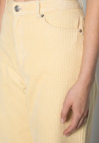 Lichtgele corduroy broek met verticale ribbels, voorzien van een voorzak en een metalen knoopdetail. Glad oppervlak met een getailleerd ontwerp.