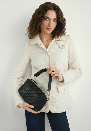 Kobieta o kręconych włosach, ubrana w jasną pikowaną kurtkę, trzymająca czarną plecioną torebkę z krótkim paskiem na jednolitym tle.