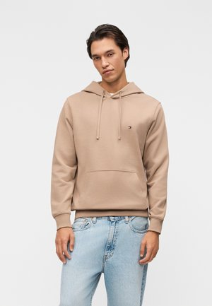 Ung mand iført beige hættetrøje med forlomme og lyseblå jeans, stående mod en ensfarvet baggrund.