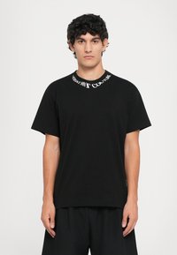 Sort t-skjorte i svart bomull med rund hals, og hvit "Versace Jeans Couture" tekst. Løs passform, korte ermer, og minimalistisk design.