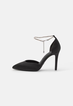 Chaussure à talon haut noire pointue avec une bride de cheville en chaîne argentée et un talon fermé sur fond blanc.