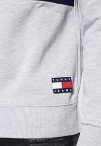 Felpa grigia con accenti blu navy, polsini a costine e un piccolo logo a patch di Tommy Jeans in colori rosso, bianco e blu. Texture morbida.