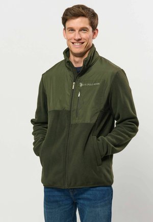 Homme souriant portant une veste polaire verte U.S. Polo Assn. avec les mains dans les poches et un jean bleu sur un fond uni.