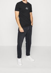 Camiseta de algodón negra con un pequeño logo blanco en el pecho, combinada con jeans gris oscuro y zapatillas blancas. El modelo está de pie con las manos en los bolsillos.