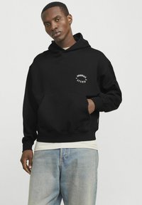 Zwarte hoodie van katoen met een kangoeroezak, voorzien van witte logo-tekst "ORIGINALS STUDIO," aansluitende manchetten en een trekkoordcapuchon.