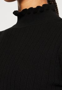Pull en tricot côtelé noir avec col montant froufroutant porté par une personne aux cheveux tressés.