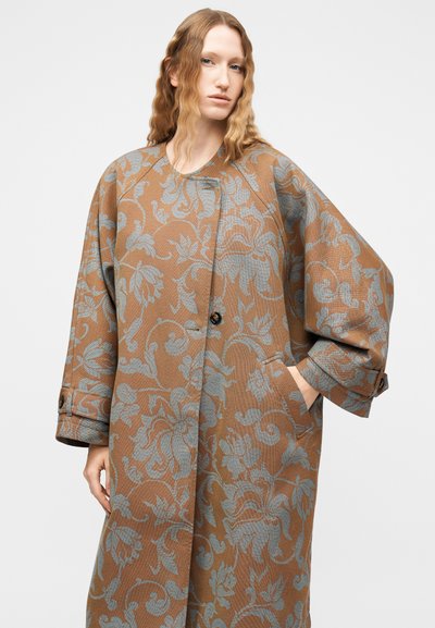Γυναίκα με μακριά κυματιστά ξανθά μαλλιά που φοράει ένα καφέ, φαρδύ παλτό με λουλουδάτο μοτίβο και μπλε-γκρι λεπτομέρειες, στέκεται μπροστά σε απλό φόντο.