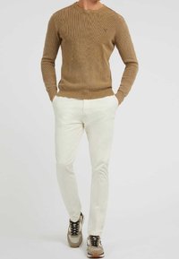 Brązowy dzianinowy sweter z prążkowaniem, okrągły dekolt, w połączeniu z jasnobeżowymi spodniami o dopasowanym kroju oraz beżowymi sneakersami z czarnymi akcentami.