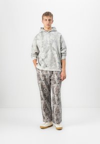 Un giovane uomo indossa un completo di felpa e pantaloni grigio camouflage abbinati, con sneakers bianche, davanti a uno sfondo bianco semplice.