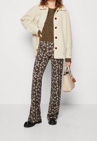 Krémová fleecová bunda s límcem a hnědými knoflíky, olivový svetr, vzorované hnědé kalhoty a světle béžová kabelka.