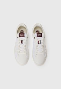 Zapatillas blancas con material suave, puntera redonda y diseño de cordones. Presenta etiquetas de logo color burdeos y acentos de tela a los lados.