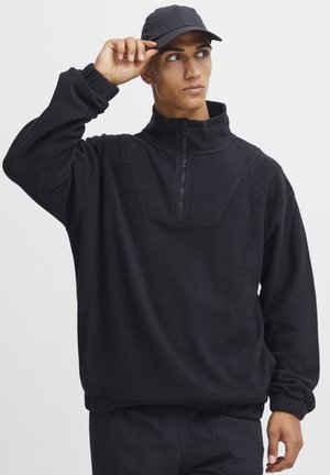 Pull noir en polaire avec un col zippé mi-hauteur, des poignets côtelés et une coupe décontractée. Présente une texture noire unie et des détails de design minimaux.