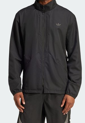 Mężczyzna w czarnej kurtce na zamek z długimi rękawami i logo Adidas na piersi, ubrany w czarne spodnie.