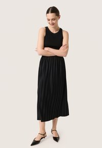 Fekete, plissé midi ruha, bordázott ujjatlan felsőrésszel, gyűrött derékkal. Fekete, hegyes orrú slingback lapos cipővel viselve.