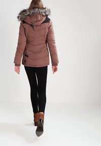 Brown puffer jacket with a faux fur hood, chevron quilting, black shoulder accents, and a side zipper pocket. Black leggings and brown heels.