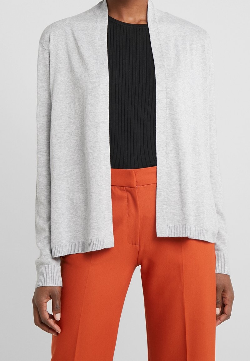 Cardigan gris clair en tricot avec devant ouvert, poignets côtelés et coupe droite, associé à un haut noir côtelé et un pantalon tailleur orange.