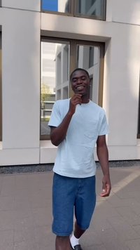Camisa de manga curta azul-claro, calções de ganga, meias brancas e chinelos pretos. De pé numa superfície pavimentada, perto de janelas grandes.