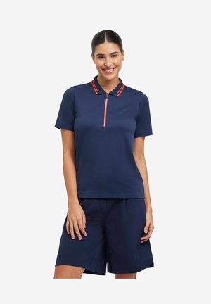 Kobieta ubrana w granatową koszulkę polo z pomarańczowym suwakiem i paskami na kołnierzyku, w granatowych szortach do kolan, uśmiecha się, trzymając ręce swobodnie wzdłuż ciała.