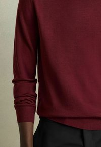 Burgundska dugmadna majica s dugim rukavima, meke teksture, s preklopljenim manžetama i opuštenim krojem, uparen s crnim hlačama.