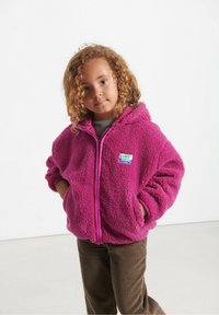 Fuzzy pink hættejakke med lynlås foran, parret med brune kordfløjl bukser. Inkluderer et lille, farverigt mærke på brystområdet.
