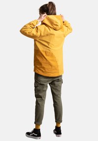 Geel hoodie-jack met een textuurafwerking, voorzien van een gebogen naaddetail, gecombineerd met olijfgroene cargobroek en zwarte sneakers.