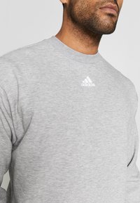 Šedá mikina s kulatým výstřihem, vyrobená z měkkého materiálu. Na hrudi se nachází malé bílé logo Adidas.