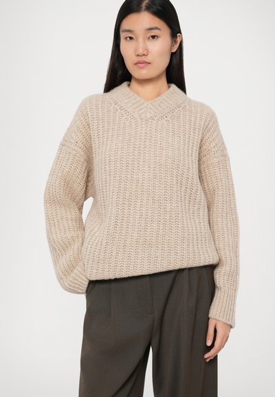 Beige gestrickter Pullover mit V-Ausschnitt und grober Textur. Der entspannte Schnitt hat überschnittene Schultern und gerippte Bündchen. Kombiniert mit dunklen Hosen.