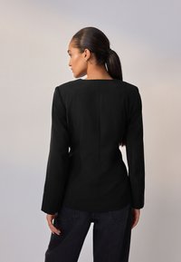 Blazer nero su misura con maniche lunghe, spalle strutturate e scollo quadrato. Abbinato a jeans in denim scuro.