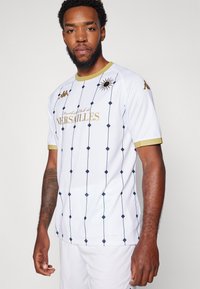 Vit kortärmad fotbollströja med blå diamantmönster och gyllene detaljer. Har klubbnamnet "Football Club de Versailles" i guld.