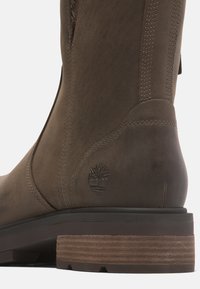 Brune lærstøvle til knæet med struktureret overflade, side-syninger og en stablet træhæl med et branded logo bagpå.