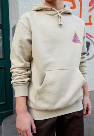 Person trägt beige Kapuzenpullover mit Fronttasche und dreieckigem Logo auf der Brust, steht neben einer mit Graffiti bedeckten Wand und einer grünen Tür.