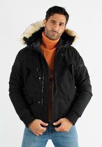 Veste noire avec capuche en fourrure, présentant un zip à l'avant, des poches latérales et des poignets côtelés. Portée sur un pull orange et un jeans bleu.