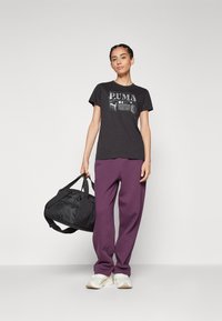T-shirt en coton noir avec un graphisme "PUMA" gris, associé à un pantalon de jogging violet. Le modèle porte un sac de sport noir et des baskets blanches.