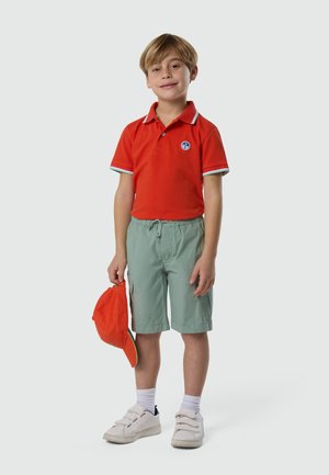 Garçon portant un polo orange et un short vert clair, tenant une casquette de baseball orange à ses côtés, debout devant un fond uni.