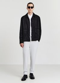 Giubbotto di suede nero con chiusura a bottoni e due tasche frontali, indossato sopra una camicia bianca e abbinato a pantaloni grigio chiaro e bott boots neri.