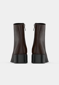 Paire de bottines marron foncé à talons carrés noirs et fermetures éclair arrière, vues de dos sur un fond blanc.