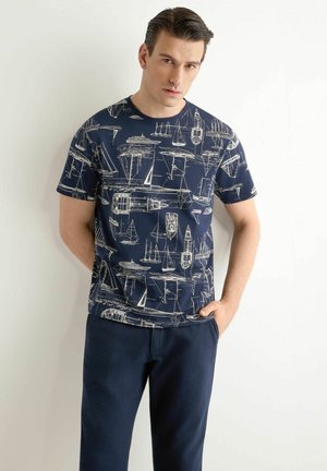 Mężczyzna w granatowej koszulce z białymi szkicami łodzi i ciemnoniebieskich spodniach, stojący na jednolitym białym tle.