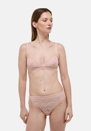Femme portant un soutien-gorge en dentelle rose pâle et une culotte assortie, debout devant un fond blanc, regardant vers sa gauche.