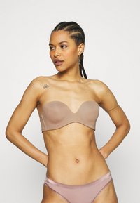 Mulher com cabelo trançado usando um sutiã nude sem alças e roupa interior correspondente, de pé com as mãos nos quadris em frente a um fundo plain.