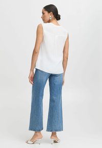 Weiße ärmellose Bluse mit hohem Kragen, kombiniert mit hellblauen weit geschnittenen Jeans. Das Outfit zeichnet sich durch einfache, glatte Texturen und minimalistisches Design aus.