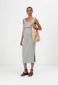 Striped, sleeveless midi dress in black and white ribbed fabric, featuring side slits, paired with a beige woven tote and black sandals.