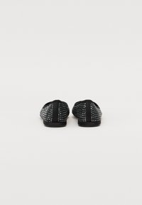 Fekete slip-on cipők texturált hálós felsőrésszel, szegecsekkel díszítve, fekete gumitalppal, hátulról nézve.