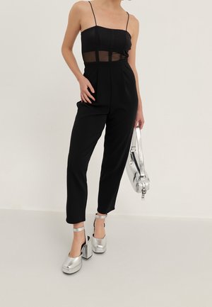 Femme portant une combinaison noire sans manches avec des panneaux transparents, tenant un sac à main argenté, et portant des talons compensés argentés avec des brides à la cheville.