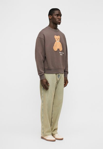 Καφέ φούτερ με ανάγλυφο σχέδιο αρκουδάκι από υλικό teddy στο μπροστινό μέρος, σε συνδυασμό με ανοιχτοπράσινο τζιν με ίσια γραμμή και παπούτσια σε κρεμ απόχρωση.