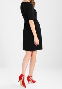 Musta lyhythihainen mekko, jossa on istuva siluetti ja taka-aukko, yhdistettynä punaisiin mokkanahkakenkiin, joissa on nilkkahihnat.