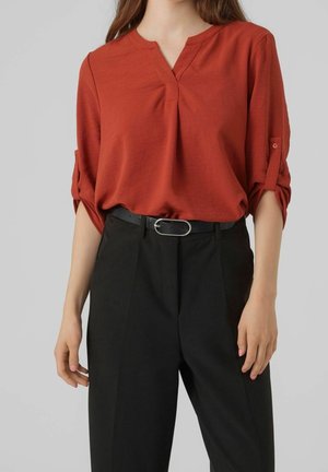 Vrouw die een roestoranje V-hals blouse draagt met opgerolde mouwen, ingestopt in een zwart broek met hoge taille, verfraaid met een zwarte riem en een zilveren gesp.