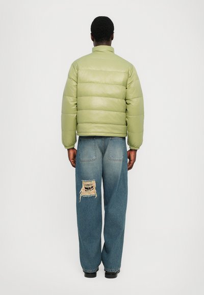 Grüne Pufferjacke mit glänzender Oberfläche und hohem Kragen. Getragen über lockeren blauen Jeans mit einem großen, ausgefransten Loch an einem Bein.