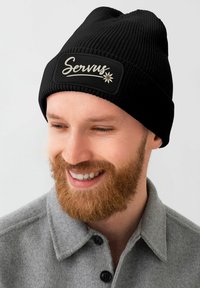 Black ribbed beanie with a rectangular patch displaying the text "Servus" in white script and a small floral design.